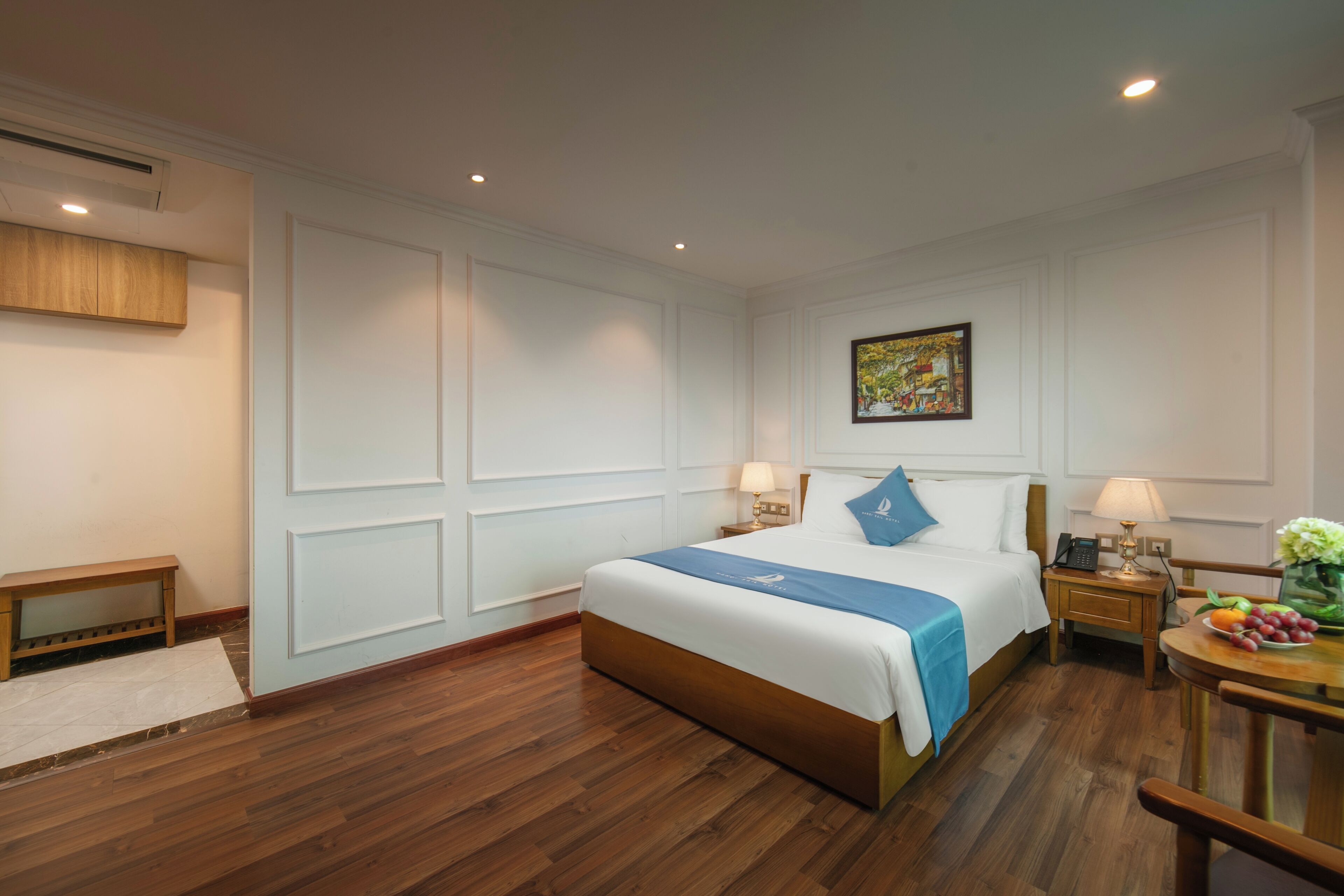 Camera d'albergo con un letto grande, comodini con lampade, una piccola panca di legno e un tavolo da pranzo con frutta.