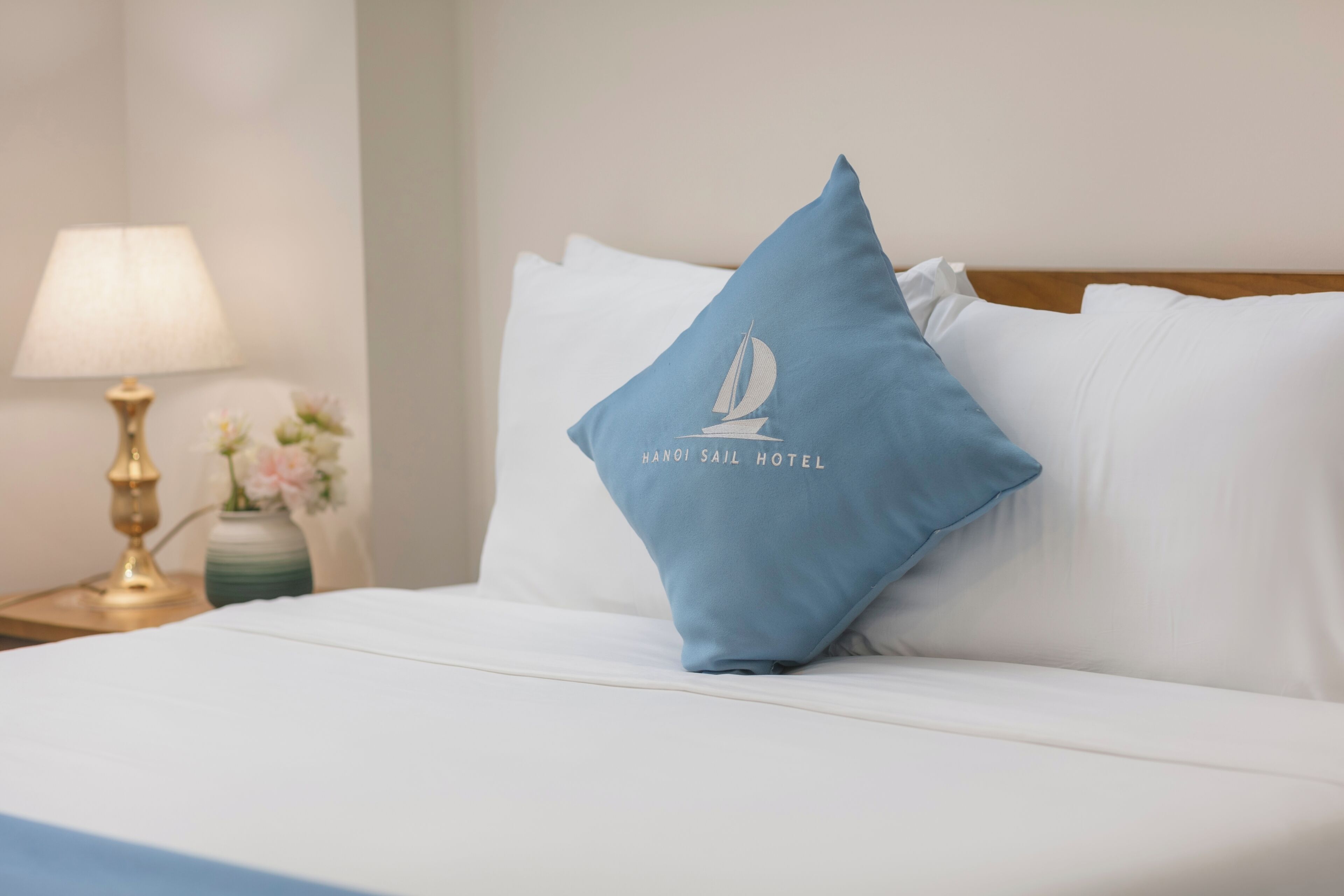 Una cama con ropa de cama blanca, un almohadón azul con el texto 'HANOI SAIL HOTEL' y una lámpara de noche, además de un jarrón con flores.