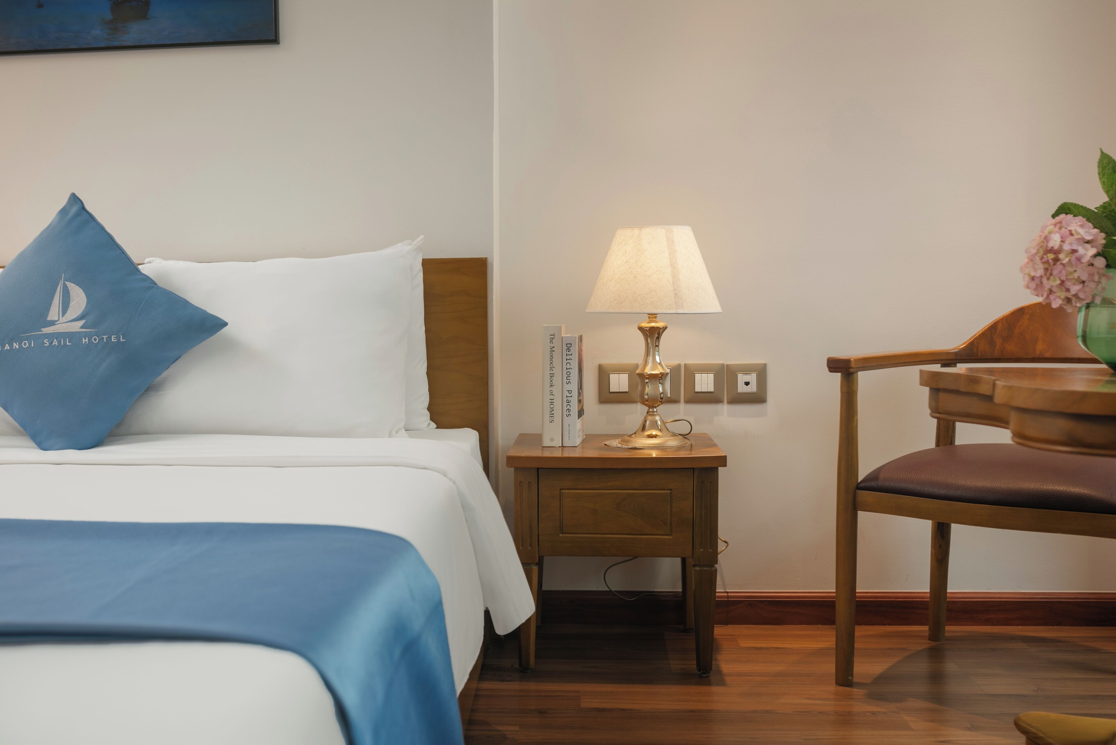 A hotel room with a bed, a nightstand with a lamp, a chair, and a wall-mounted book.