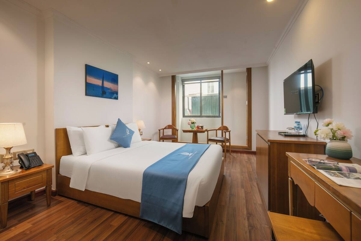 Habitación de hotel con cama, mesita de noche, lámpara, teléfono, escritorio, silla, televisor y ventana.