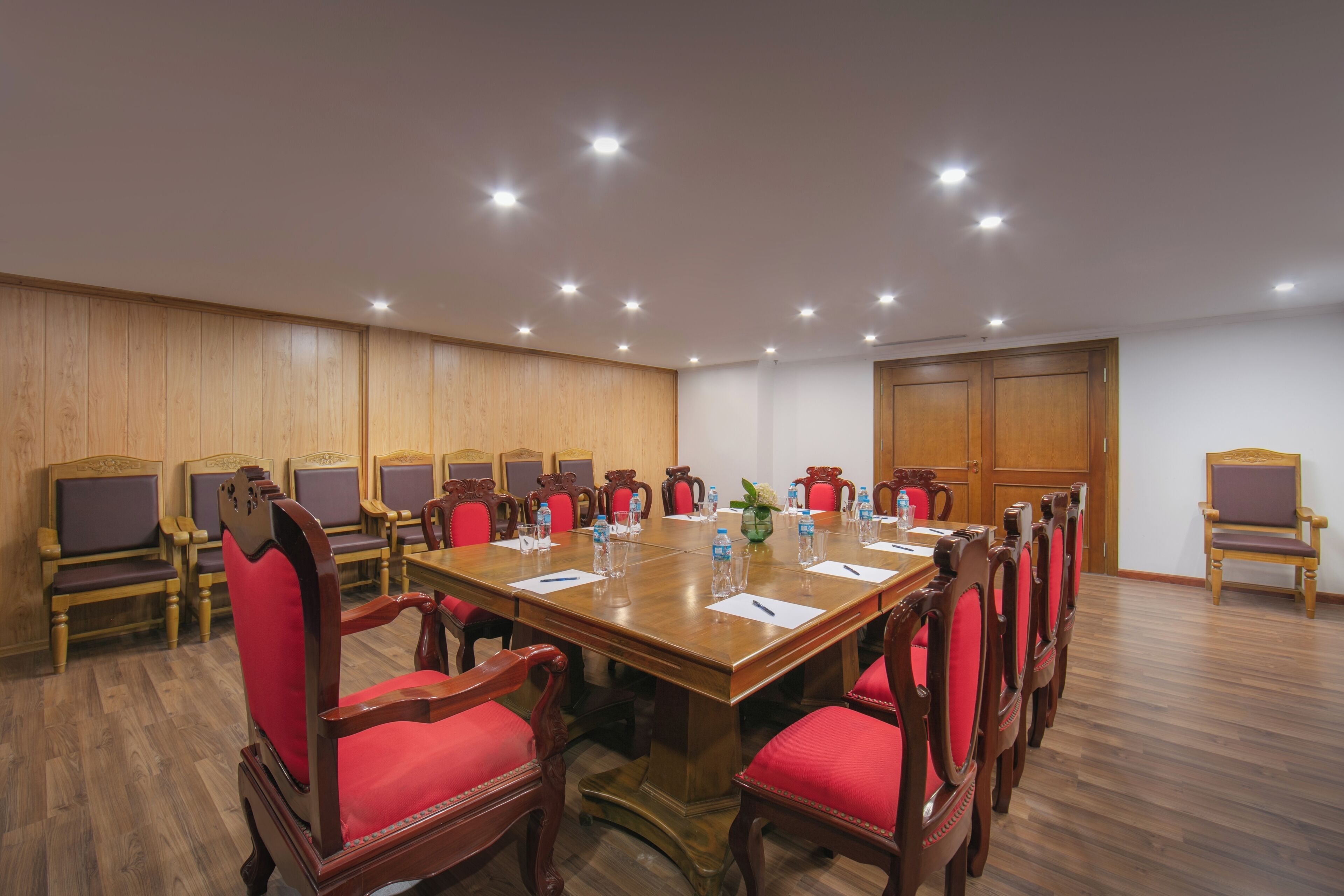 Une salle de conférence avec une longue table en bois, des chaises rembourrées de couleur rouge et des spots encastrés au plafond.