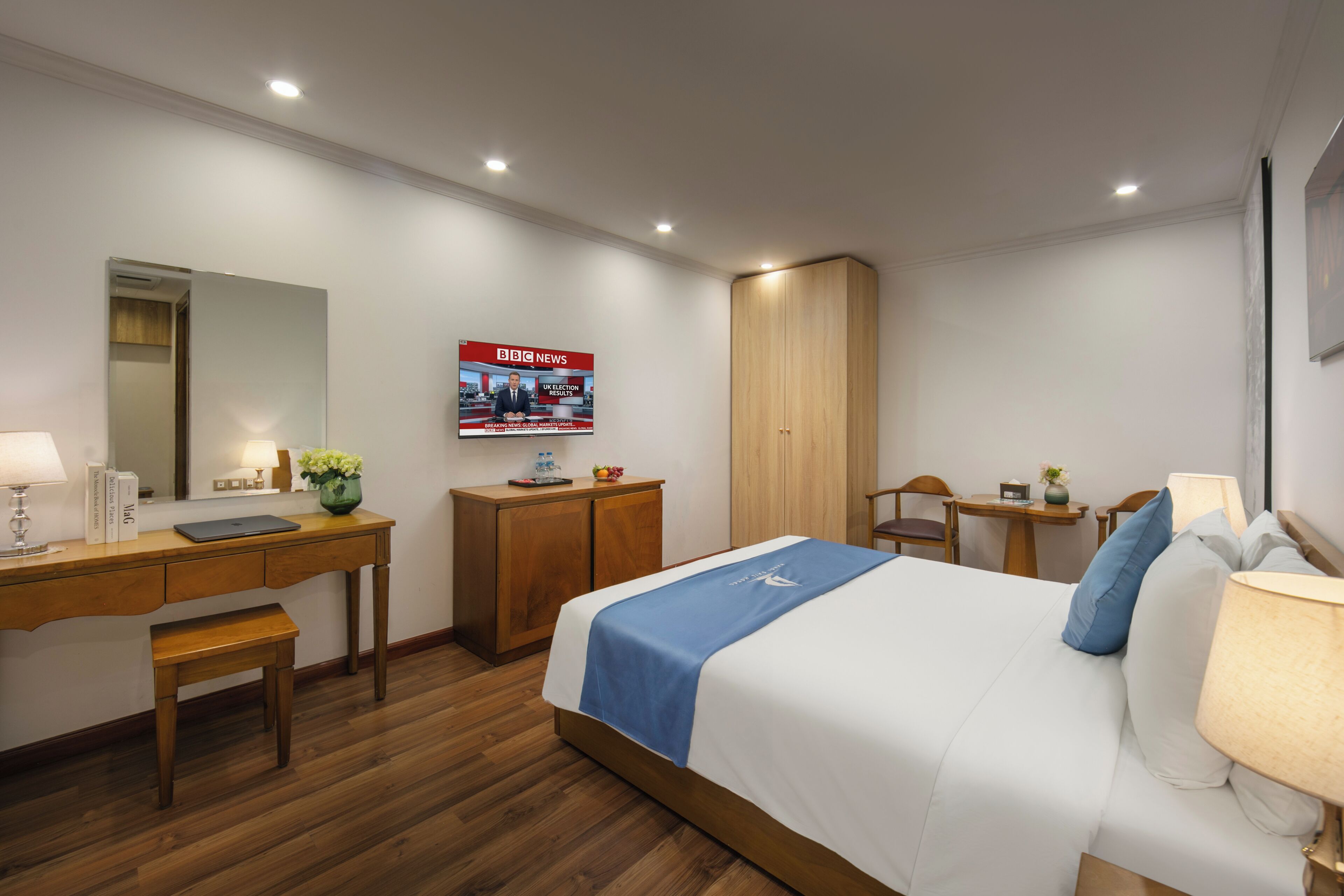 A hotel room with a bed, desk, chair, wardrobe, and a television displaying news.