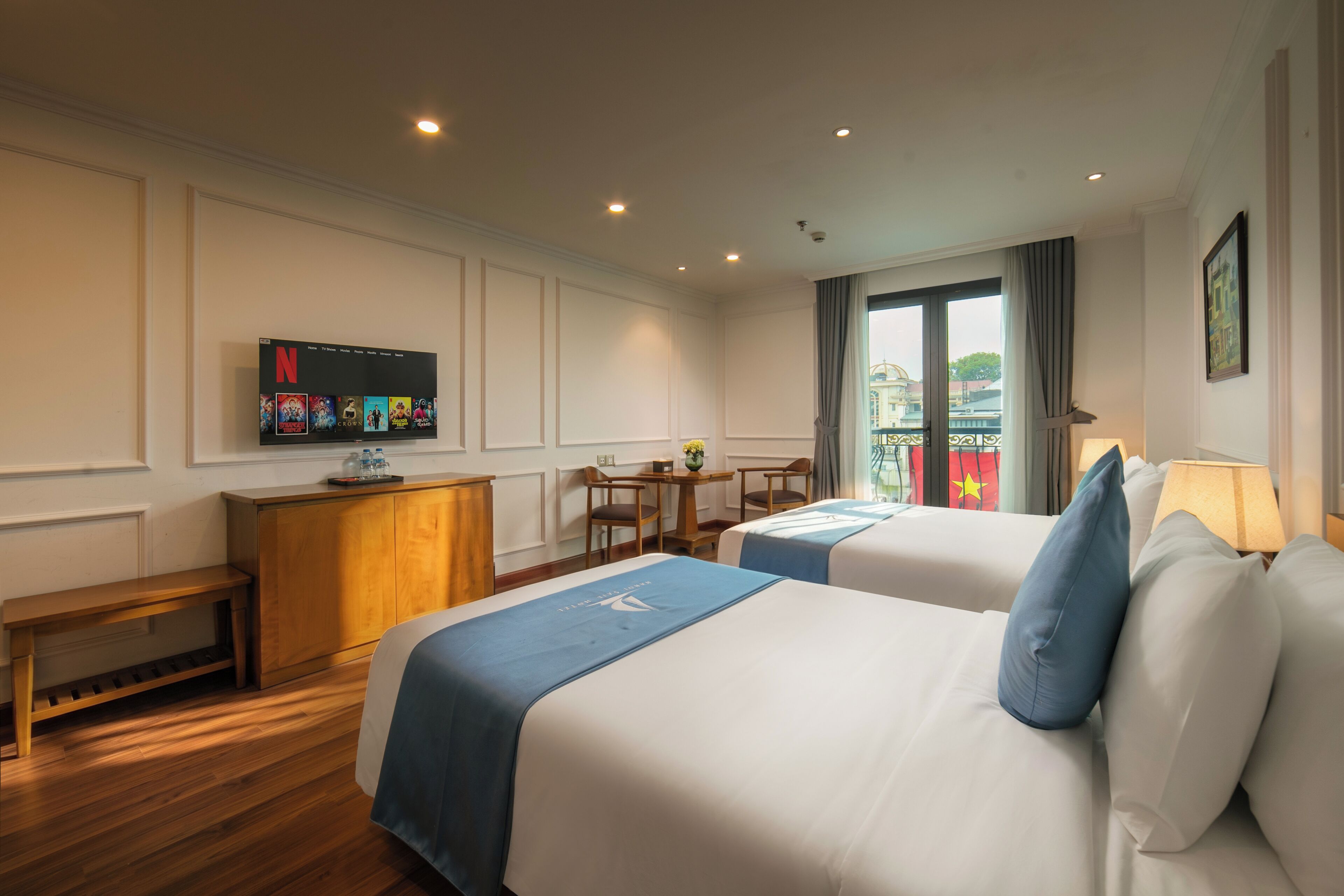Habitación de hotel con dos camas, un televisor montado en la pared, un escritorio con sillas y vistas a un edificio.