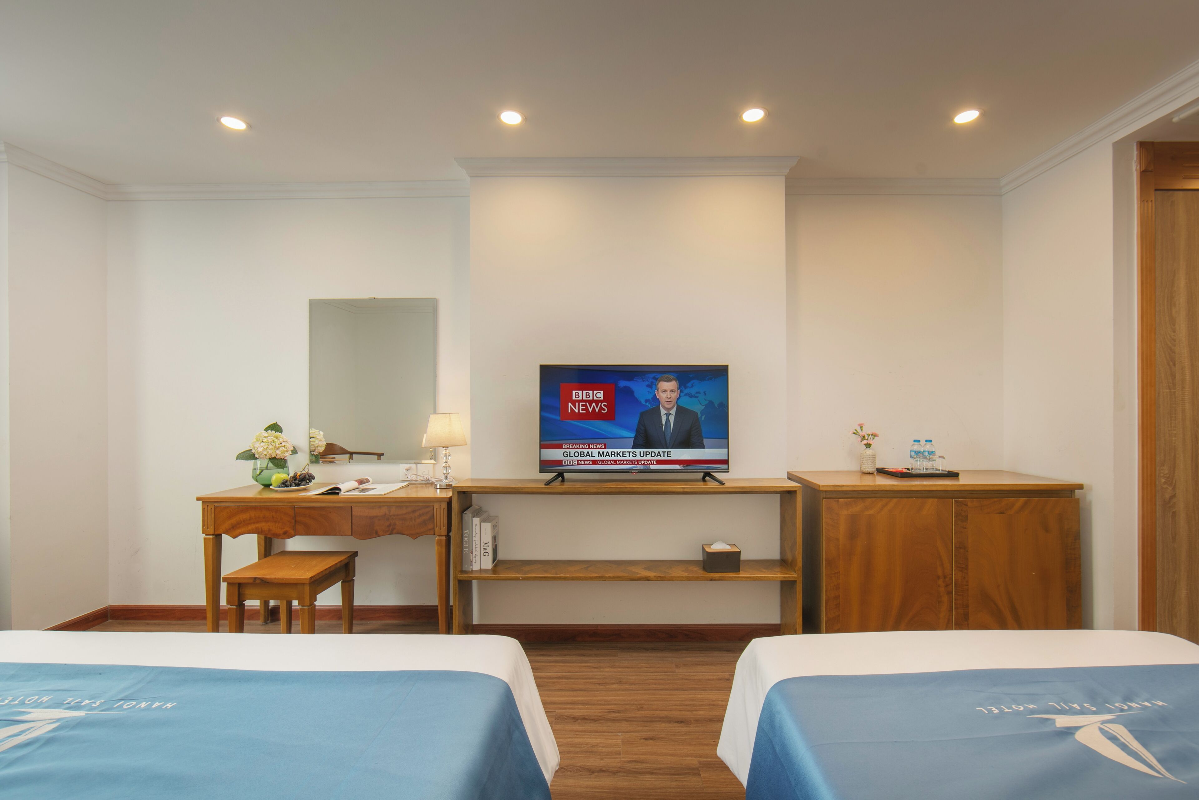 A hotel room with two beds, a desk, a television displaying the BBC News, and a wooden cabinet.