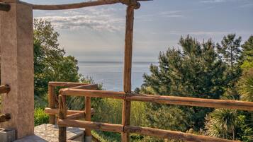 A wooden balcony with a stone floor, overlooking a coastal landscape with trees and the sea.