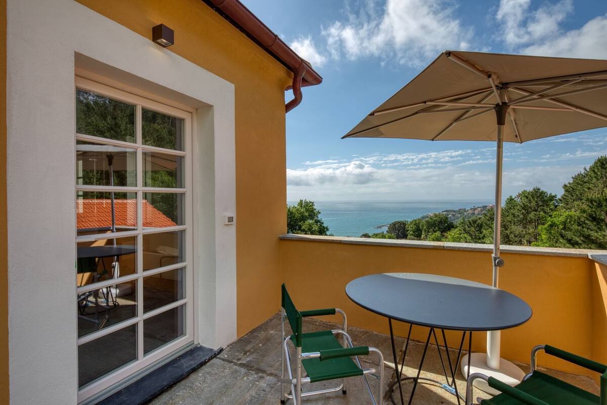 Un balcon avec une table et des chaises, un grand parasol et une vue sur la mer et les collines.