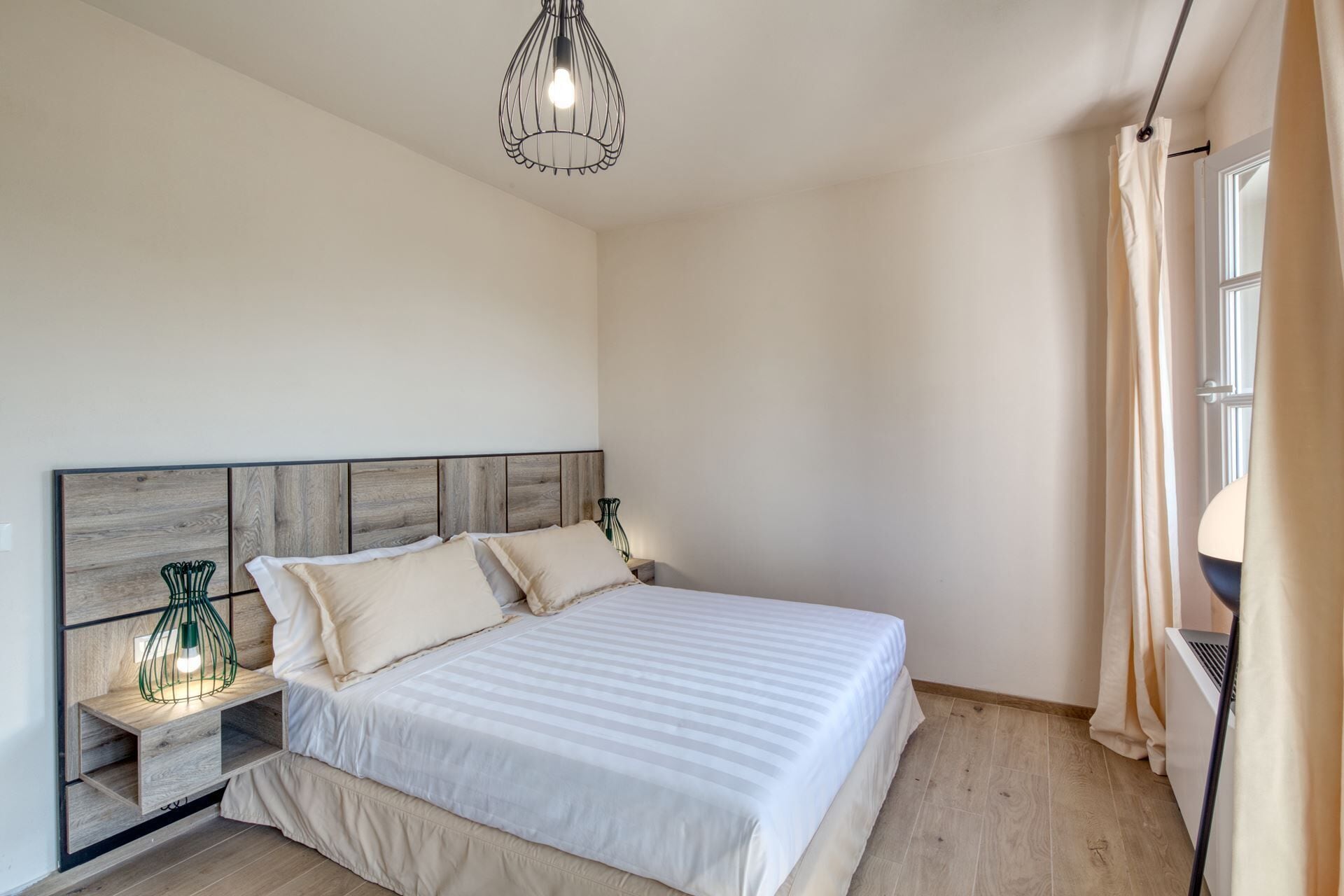 A bedroom with a wooden headboard, a bed with white bedding, a nightstand with a green lamp, and a window with beige curtains.