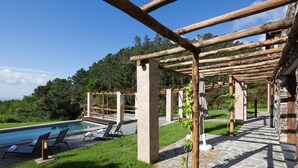 Un área junto a la piscina con pérgolas de madera, sillones de descanso y un camino de piedra.