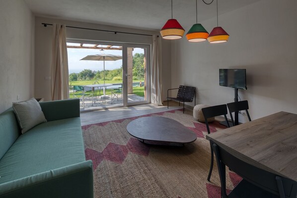 Una sala moderna con un sofá verde, una mesa de centro redonda y una mesa de comedor de madera. Hay un televisor sobre un mueble bajo y se ve un jardín a través de la puerta de cristal.