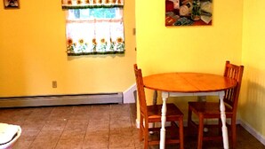 A dining area with a wooden table and chairs, a window with a floral curtain, and a painting on the wall. - Large 1 bed w/ private deck (Portland)