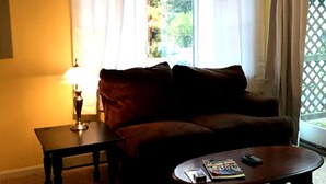 A living room with a brown sofa, a small table, and a coffee table. There is a window with curtains, a lamp, and a view of greenery outside. - Large 1 bed w/ private deck (Portland)