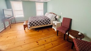 A bedroom with a wooden floor, a bed with a patterned bedspread, a nightstand, a chair, and a desk with a lamp. - Studio Apartment near downtown Walkable! (Portland)