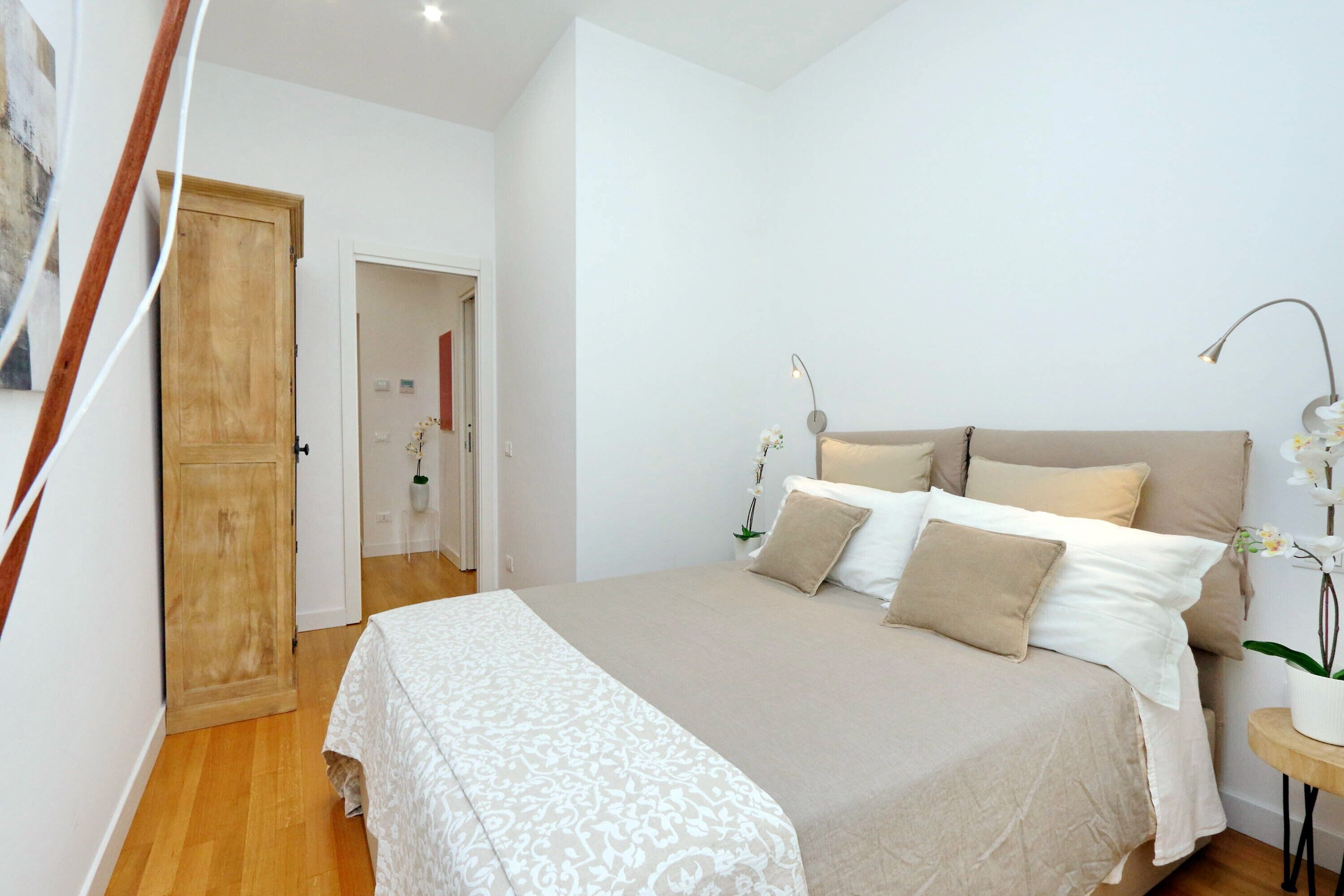A bedroom with a large bed, a wooden door, a small table with a lamp, and a potted plant.