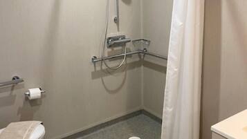A small, enclosed shower area with a white showerhead, a handheld showerhead, and a shower curtain. There is a toilet with a towel on it and a small sink with a mirror.
