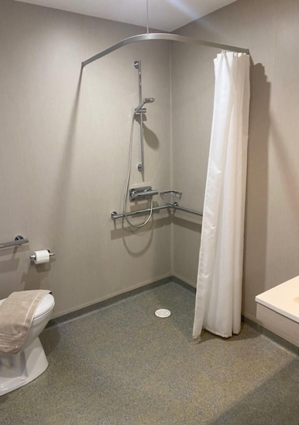 Un espacio de ducha pequeño y cerrado con ducha blanca, ducha de mano y cortina de ducha. Hay un inodoro con una toalla y un lavamanos pequeño con espejo.