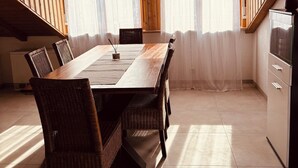 Ruokailutilassa on puinen pöytä ja punottuista materiaaleista valmistetut tuolit, seinällä on kello ja keittiöalueella on jääkaappi ja kaapit.