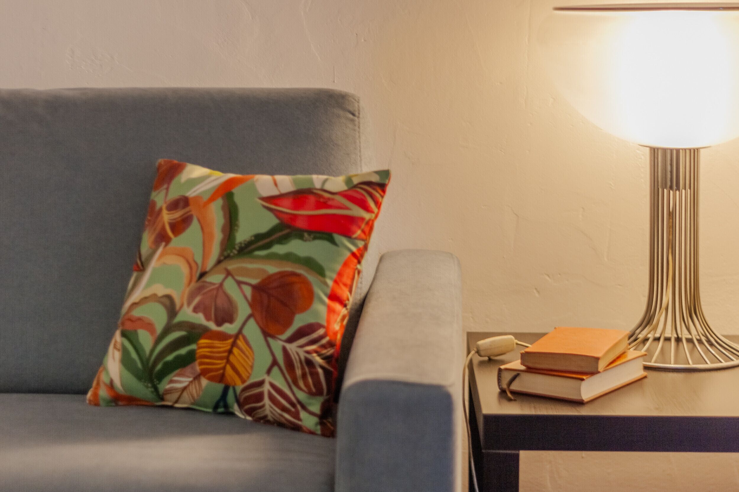 Un canapé agrémenté d’un coussin coloré, une lampe et une pile de livres posés sur une table d’appoint.