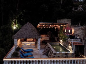 A thatched-roof structure with outdoor seating and a pool area, surrounded by stone walls and vegetation.