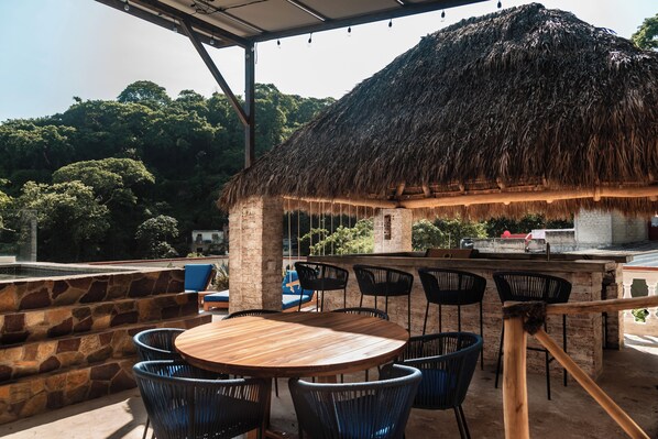 An outdoor seating area with a round wooden table and black chairs, a thatched-roof structure, and a view of a forested hillside.
