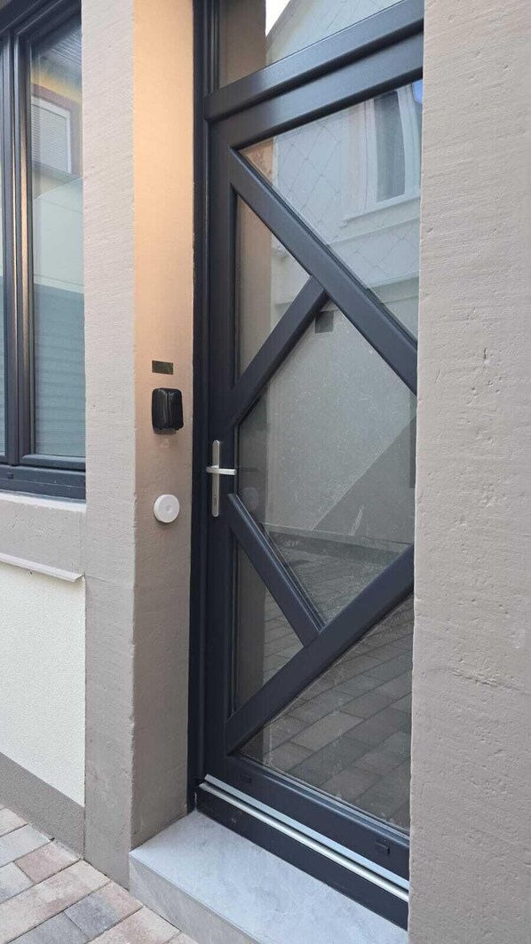 Una puerta moderna con un panel de vidrio y una manija, ubicada en un edificio con una pared exterior sencilla y una ventana a la izquierda.