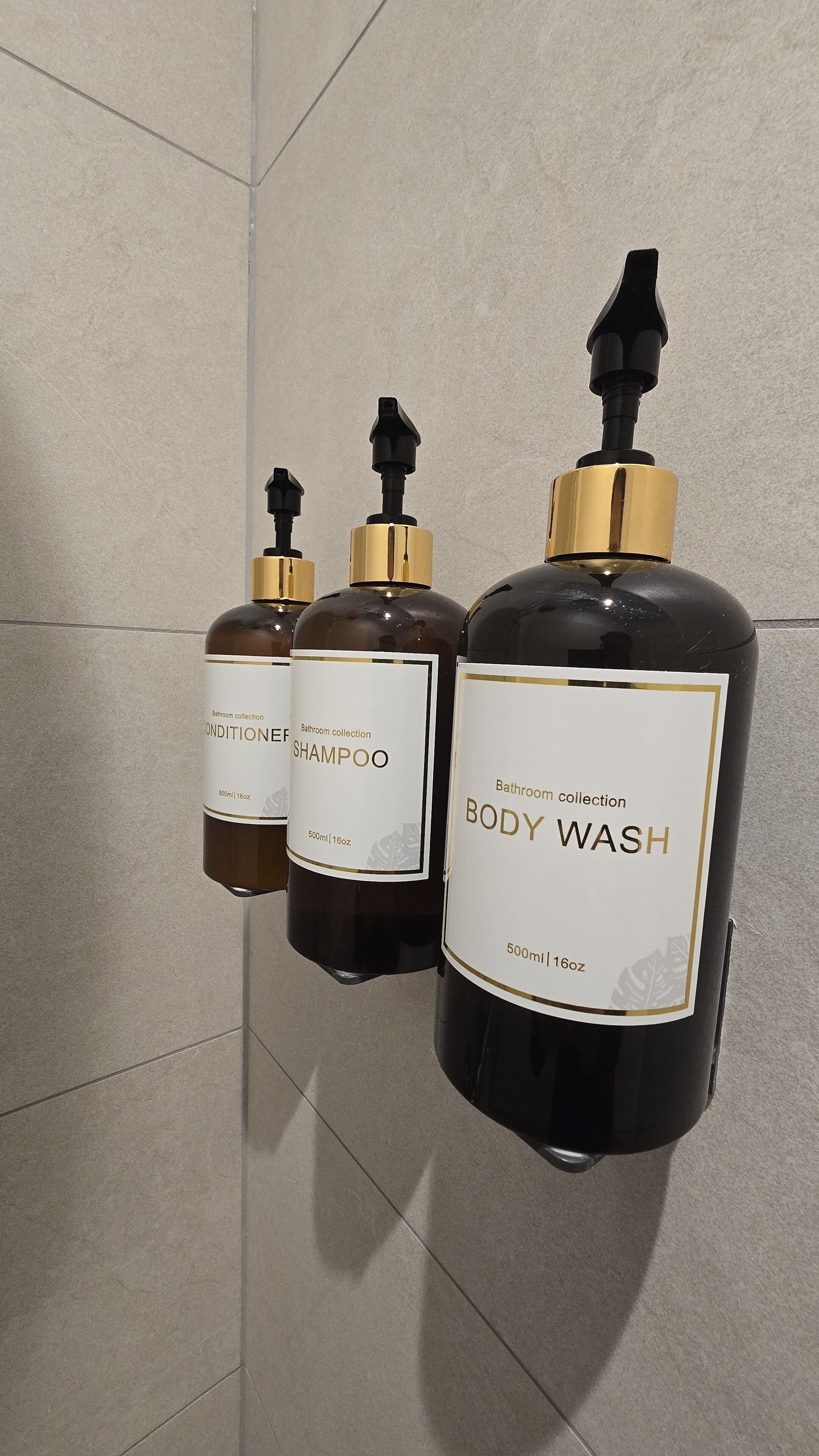 Three bottles of bathroom collection body wash and shampoo, each with a 500ml/16oz capacity, mounted on a tiled wall.