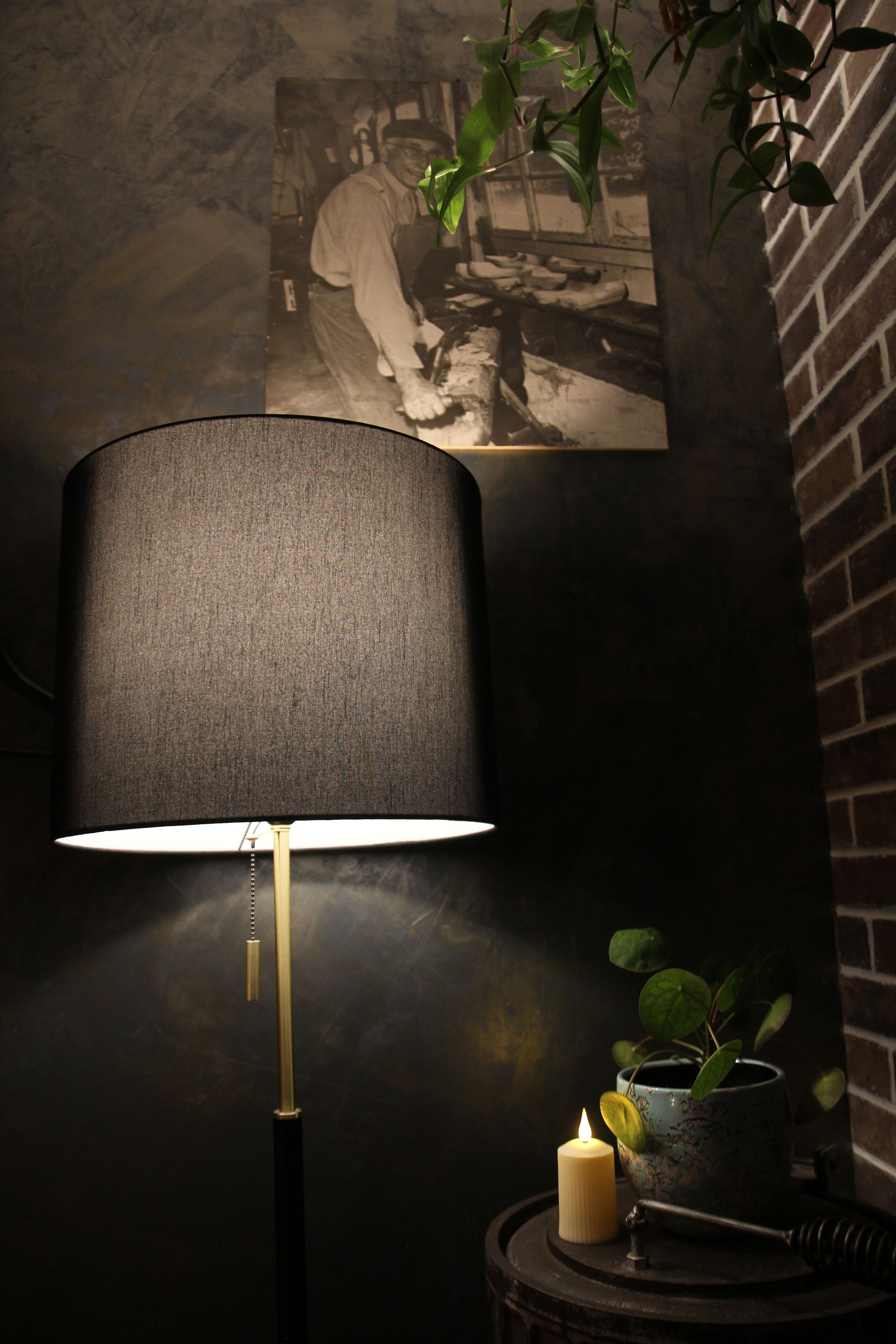 Eine Lampe mit dunklem Schirm, eine brennende Kerze und ein kleiner Topfpflanze auf einem Tisch.