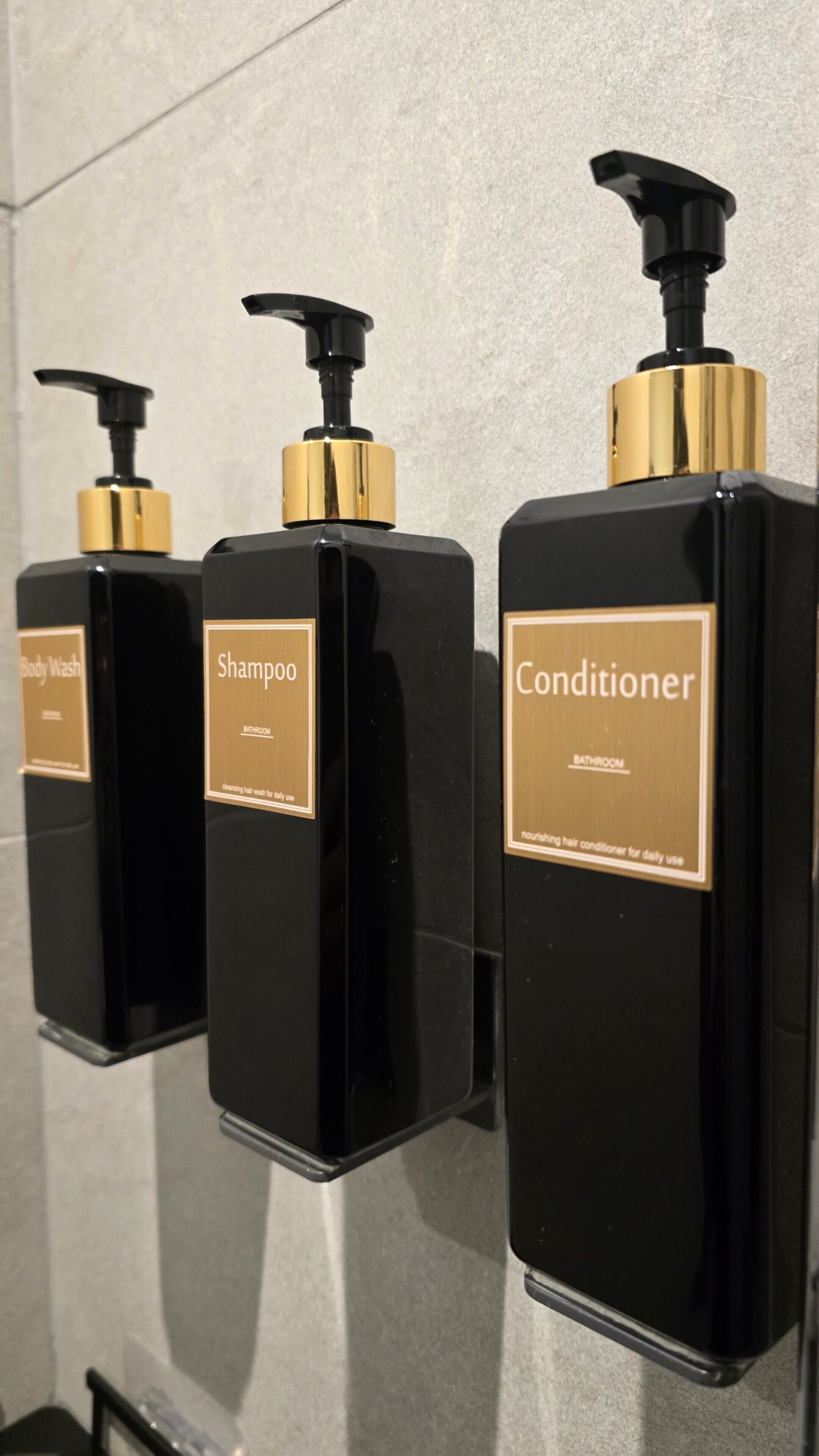 Drie zwarte flessen met gouden details, gelabeld 'Body Wash', 'Shampoo' en 'Conditioner', in een badkamer.