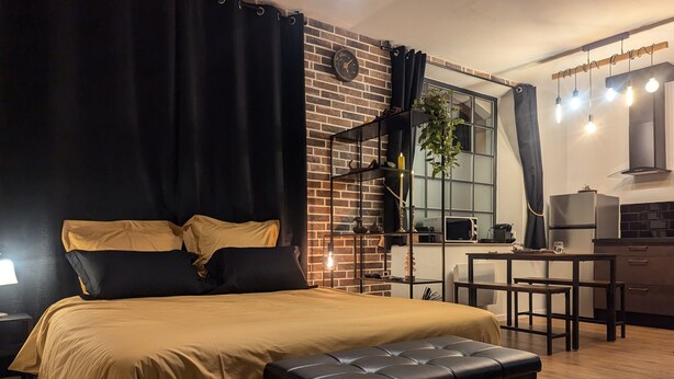 A bedroom with a bed, a bench, a desk, and a kitchen area.