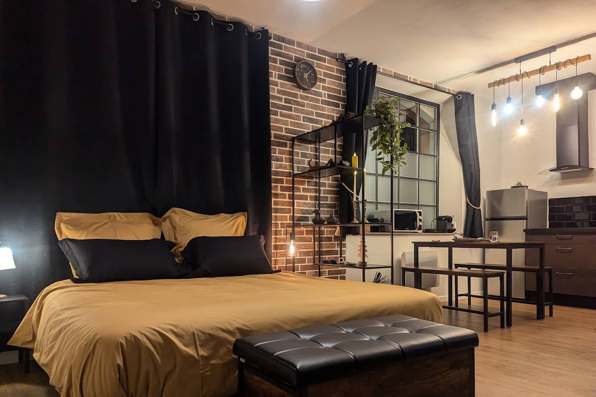 A bedroom with a bed, a bench, a desk, and a kitchen area.