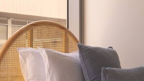 A bedroom with a wicker headboard, white bedding, and a view of a balcony with plants and clothes hanging. - Coastal Retreat Steps to Beach (Port Melbourne)