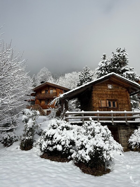 A wooden cabin with a stone foundation, surrounded by snow-covered trees and a wooden bridge.