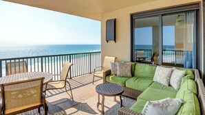 Ein Balkon mit Blick auf das Meer, ausgestattet mit Korbstühlen, einem runden Tisch und einem grünen Sofa mit gemusterten Kissen.