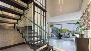 A modern interior with a glass staircase, a living area with a view, and a decorative wall piece. - Gayle Villa - Stunning Modern Masterpiece with Breathtaking Views (Los Angeles)