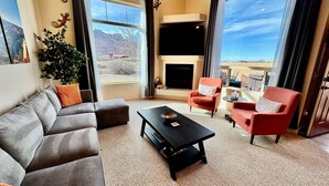 A modern living room with a grey sectional sofa, a black coffee table, and two red armchairs. A large window offers a view of a mountainous landscape.