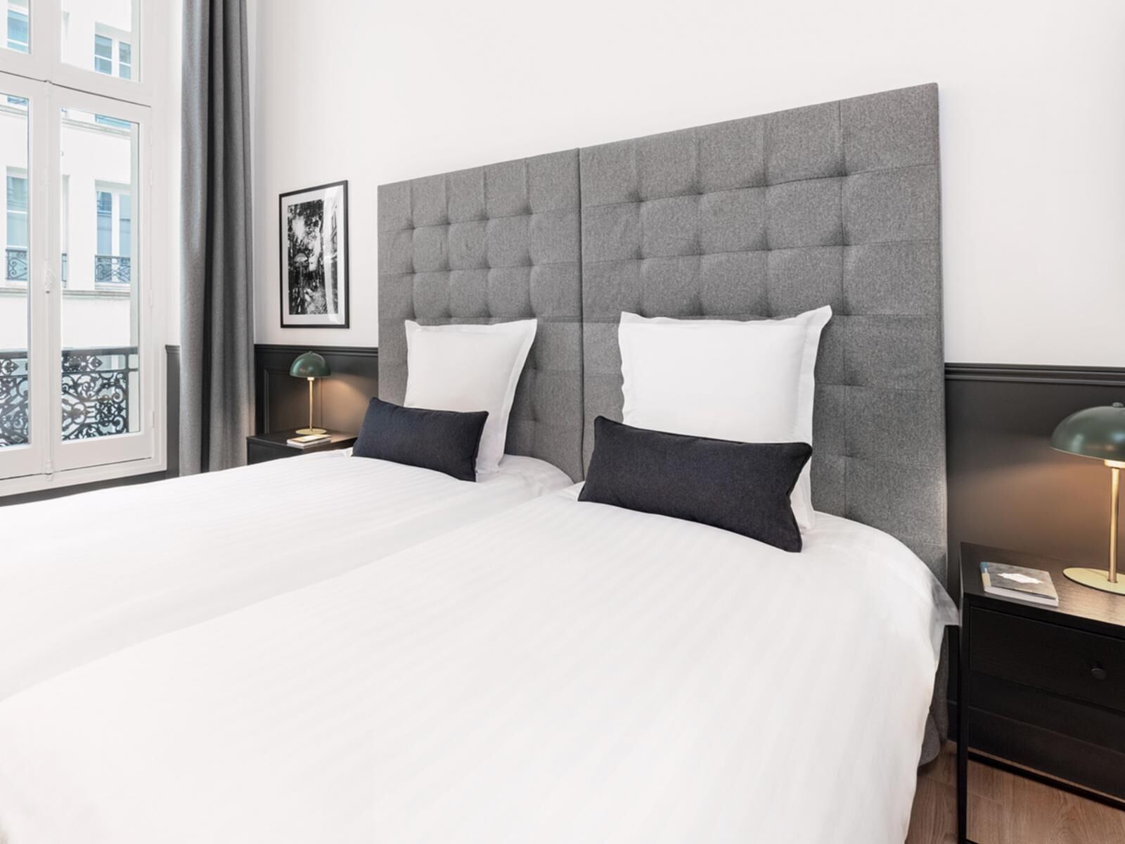 Une chambre d’hôtel moderne avec un grand lit, une tête de lit rembourrée de couleur grise, du linge de lit blanc et deux tables de chevet avec des lampes.