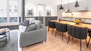 Uma sala de estar moderna com um sofá cinza, uma mesa de jantar com cadeiras, e uma área de cozinha com armários e uma fruteira.