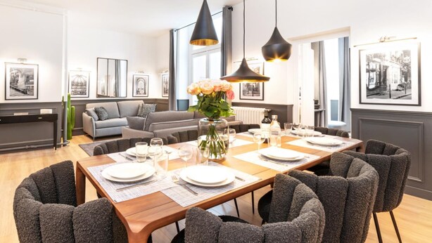 Uma sala de jantar com uma mesa de madeira posta para seis pessoas, rodeada por seis cadeiras cinzas estofadas.