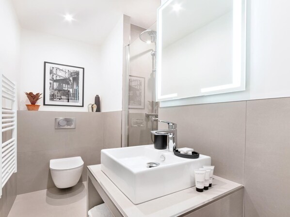 A modern bathroom with a white sink, toilet, and shower. There is a framed picture on the wall and a potted plant on the counter.