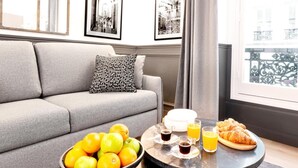 Uma sala de estar moderna com um sofá cinzento, um travesseiro com padrão preto e branco, uma tigela de frutas, uma mesa de centro com itens de café da manhã, e uma janela com vista para uma varanda.