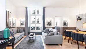 Uma sala de estar moderna com um sofá cinzento, uma mesa de centro e um espaço de jantar com cadeiras. Há quadros nas paredes e uma janela grande com cortinas.