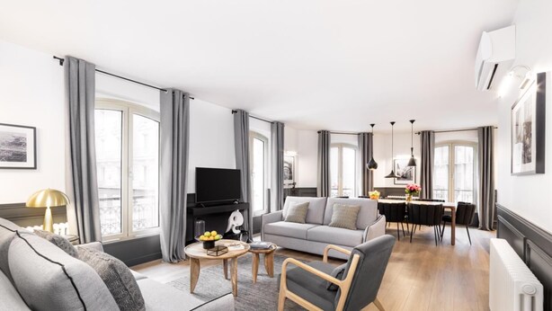 Ein modernes Wohnzimmer mit einer grauen Couch, einem kleinen Couchtisch und einem Fernseher. Im Hintergrund befindet sich ein Essbereich mit Tisch und Stühlen.