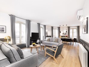 Un salon moderne avec un canapé gris, une petite table basse et un téléviseur. On aperçoit à l’arrière-plan un coin repas avec une table et des chaises.