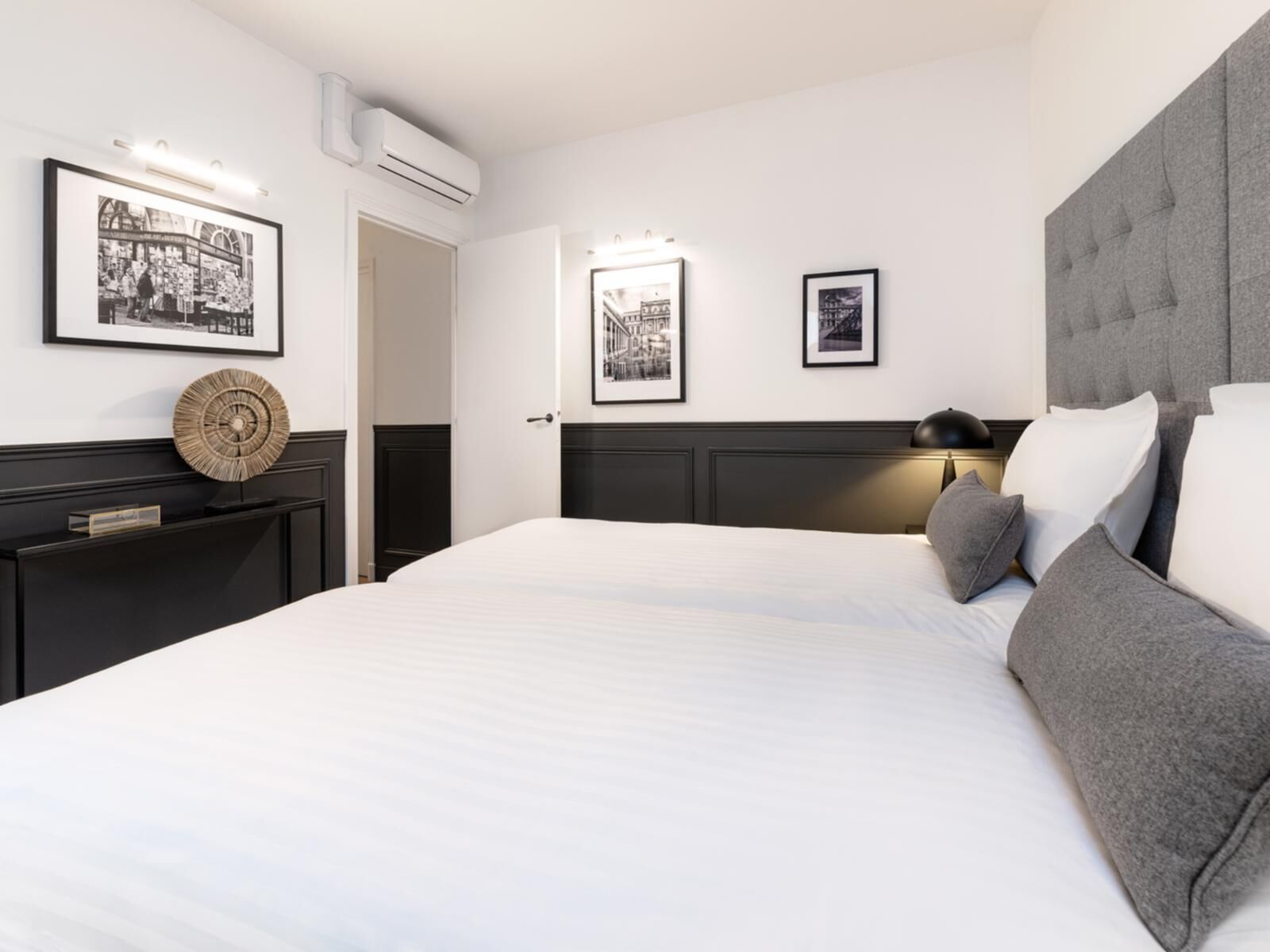 Een moderne hotelkamer met een groot bed, een donker hoofdeinde en twee nachtkastjes. Er hangen ingelijste foto’s aan de muur en boven het bed bevindt zich een airconditioner.