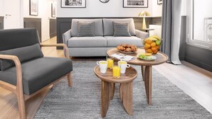 Ein modernes Wohnzimmer mit einem grauen Sofa, einem Holzstuhl, einem Couchtisch mit Frühstücksgebäck und gerahmten Kunstwerken an der Wand.