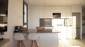 Een moderne keuken met witte kasten, een roestvrijstalen fornuis en een inbouwoven. Er staan twee witte barkrukken bij een klein eiland met een mandje.