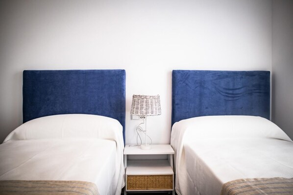 A double bed room with two blue headboards, a small bedside table with a lamp, and a wall-mounted shelf.