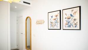 Un pasillo con un espejo grande, dos cuadros enmarcados con motivos florales y una puerta que da a otra habitación.