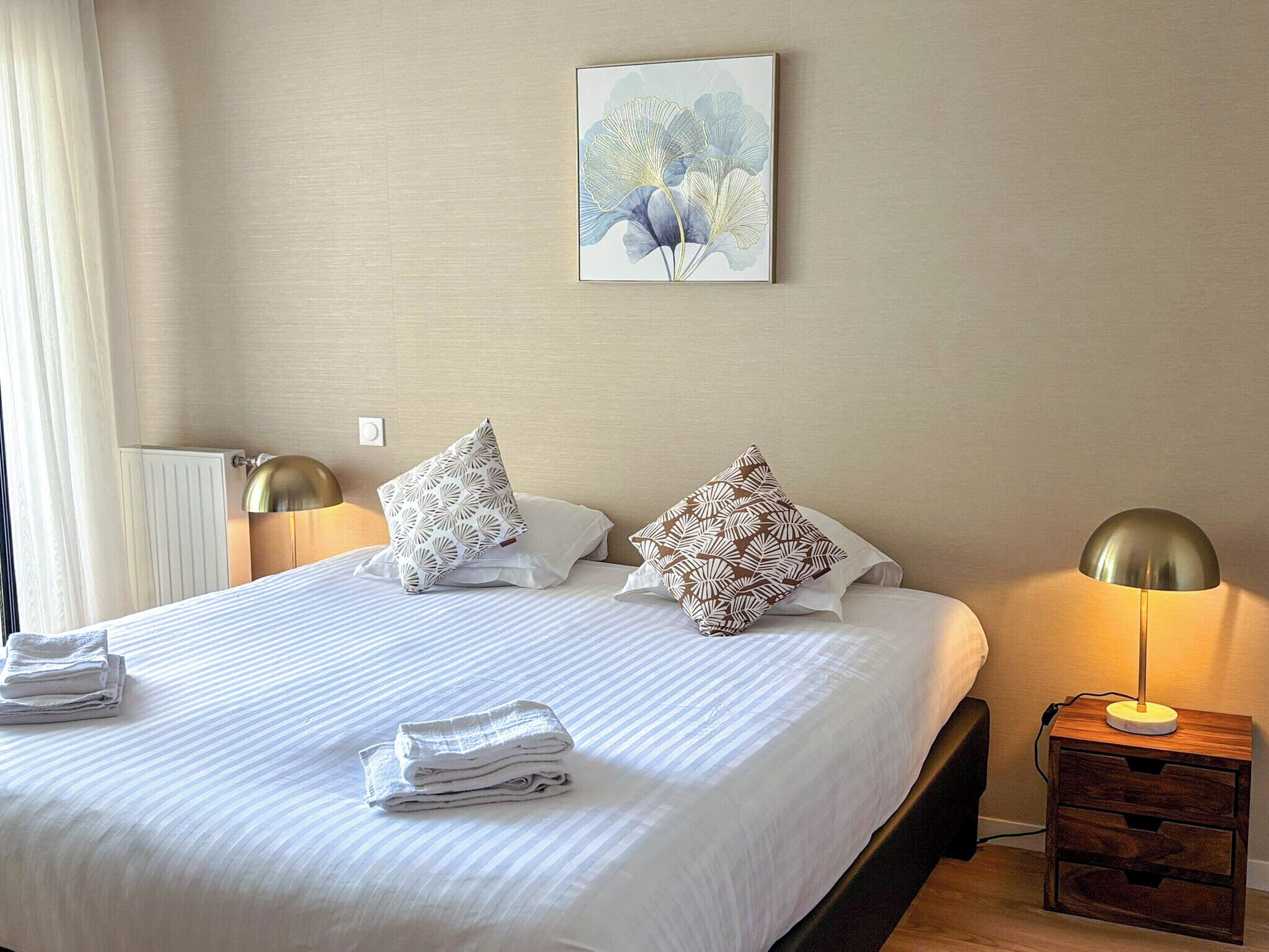 A neatly made bed with white linens and two decorative pillows, a bedside table with a lamp, and a wall-mounted artwork.