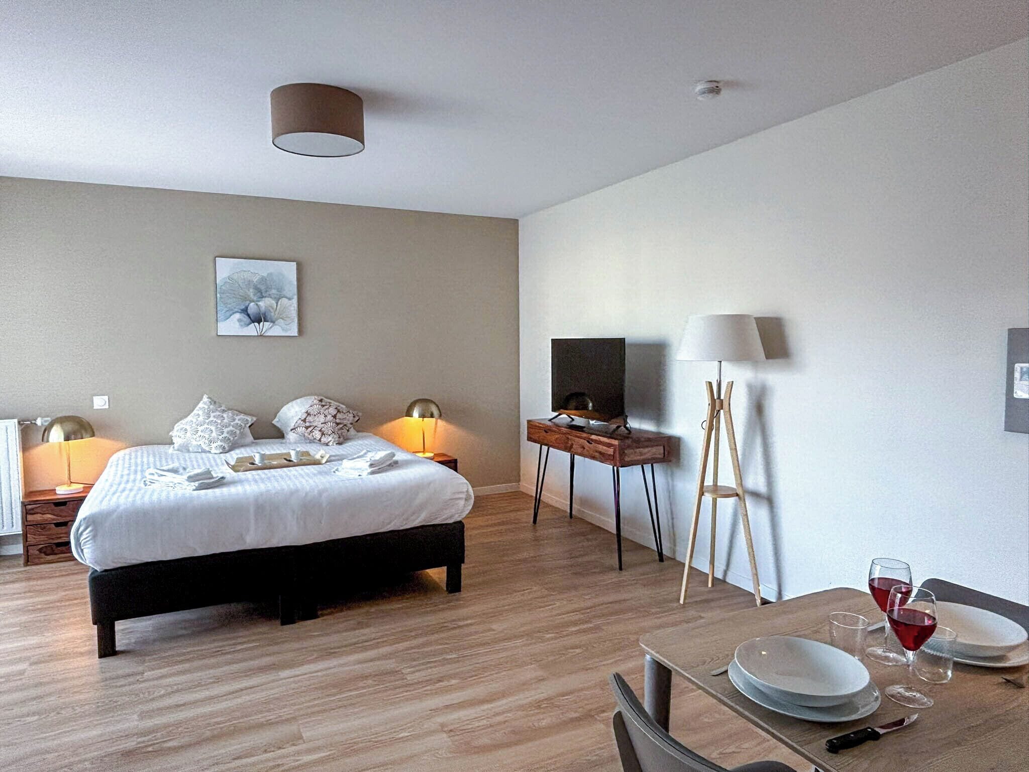 Ein modernes Hotelzimmer mit einem Bett, Nachttischlampen, einem Fernseher, einem Gemälde und einer Essecke mit Tellern und Gläsern.