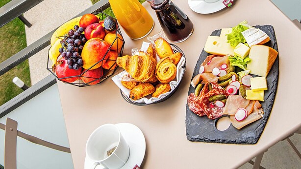 Ein Balkontisch mit Frühstücksangebot, darunter Früchte, Gebäck, Käse und Fleisch, mit Blick auf eine Terrasse und Sitzmöglichkeiten im Freien.
